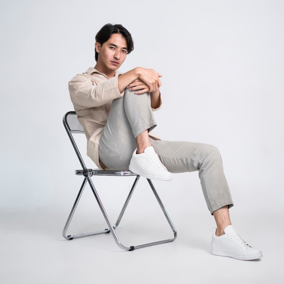 Man sitting on a chair wearing a beige shirt and light gray pants against a white background