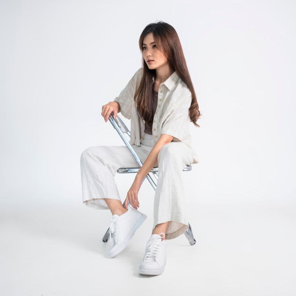 Woman sitting on a chair wearing a light-colored outfit with white sneakers on a white background
