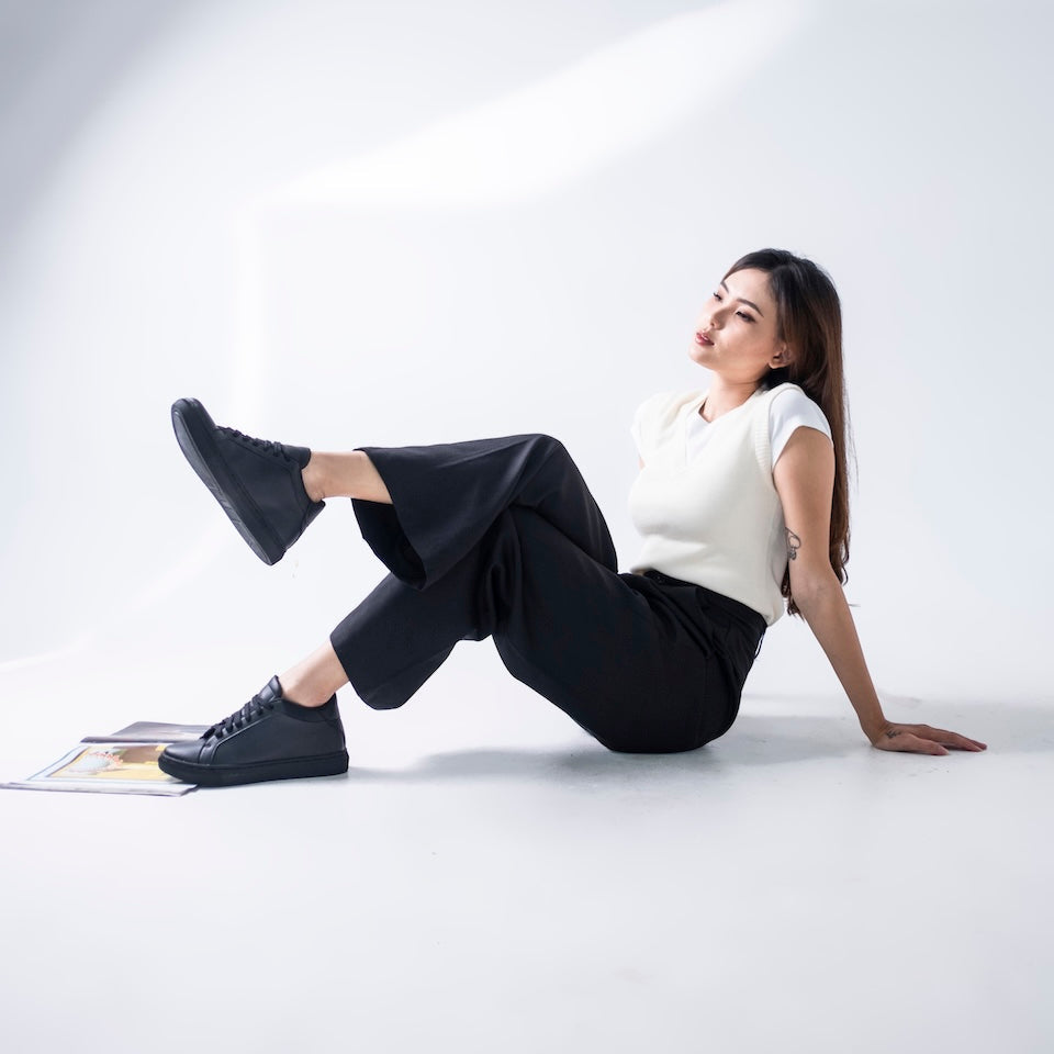 Woman sitting on a white floor wearing a white top and black pants with black shoes.
